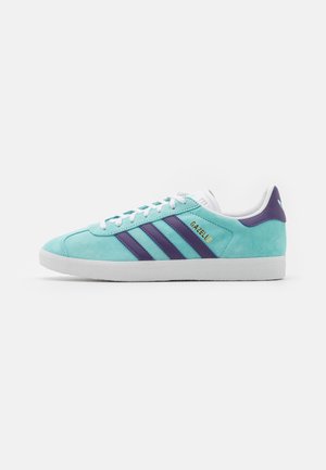 Baskets Adidas Gazelle bleu clair avec des bandes bleu foncé, lacets blancs et semelle grise, vues de profil sur un fond uni.