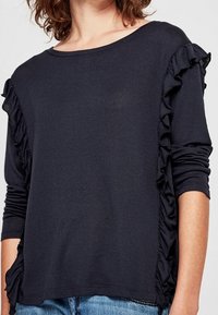 Marinblå långärmad topp med volangdetaljer längs sidorna. Tillverkad av mjukt tyg, har en bred halsringning och avslappnad passform.