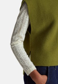 Osoba nosící bílý krajkový top s dlouhým rukávem pod bezrukávým olivově zeleným pleteným světrem, rukou v tmavě modré džínové kapse.