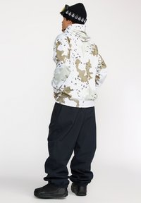 Sudadera de camuflaje en blanco, beige y negro, con un cuello alto y un bolsillo tipo canguro. Combinada con pantalones negros de corte holgado y botas.
