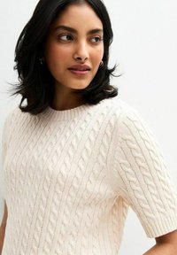 Sweter z warkoczy w jasnym kremowym kolorze, z krótkimi rękawami, okrągłym dekoltem i teksturowanym wzorem na całej powierzchni. Miękka, dziana tkanina.