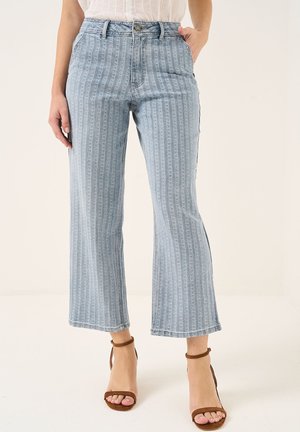 Kvinde iført lyseblå stribede cropped jeans og brune ankelremssko, stående med den ene hånd i lommen.
