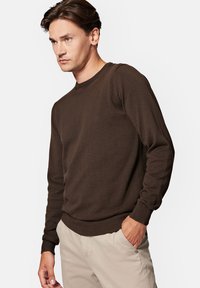 Brązowy sweter dzianinowy z długimi rękawami, okrągłym dekoltem, prążkowanymi mankietami i dołem. Małe logo na piersi. Gładka faktura.