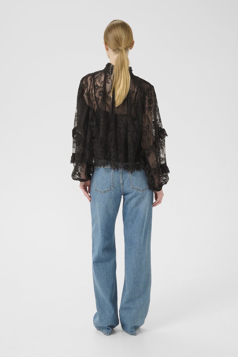 Schwarze Spitzenbluse mit langen Puffärmeln, durchscheinendem Material und gewellten Kanten, kombiniert mit hochsitzenden hellblauen Jeans.