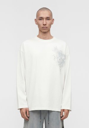 Άνδρας με ξυρισμένο κεφάλι φοράει λευκό πουκάμισο με μακριά μανίκια και διακριτικό λουλουδάτο σχέδιο στο αριστερό στήθος και γκρι παντελόνι, στέκεται μπροστά σε απλό φόντο.