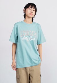 Világoskék Nike póló, laza szabással, fehér és barack színű grafikával, amelyen az "Athletic Dept" felirat látható, valamint egy apró logó az alján.