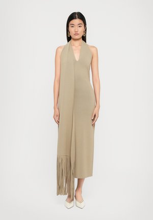 Woman wearing a sleeveless beige midi dress with deep V-neck and long fringe scarf detail, paired with white pointed shoes and gold earrings.