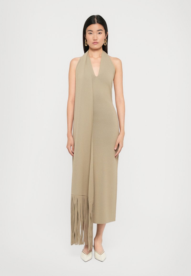 Woman wearing a sleeveless beige midi dress with deep V-neck and long fringe scarf detail, paired with white pointed shoes and gold earrings.