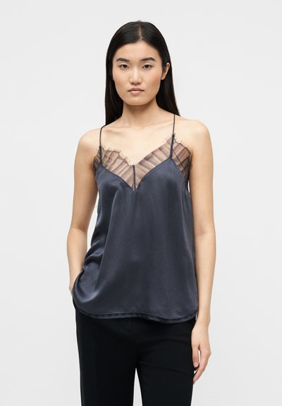 Γυναίκα με μακριά σκούρα μαλλιά, ντυμένη με ένα αμάνικο σκούρο γκρι σατέν τοπ με διαφανές βολάν στο ντεκολτέ και μαύρο παντελόνι, στέκεται μπροστά σε απλό φόντο.