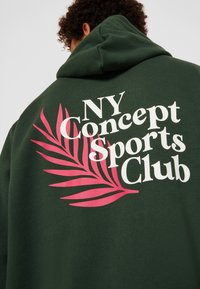 Sudadera verde con un gran gráfico en la parte posterior que presenta texto en blanco y rosa que dice "NY Concept Sports Club" y un diseño de hoja.