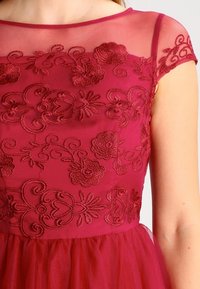 Burgundská šaty s průhledným tělem, které je zdobené vy�šitými květinovými vzory. Krátké rukávy a vrstvená tylová sukně vytvářejí texturovaný vzhled.