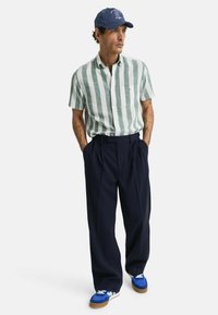 Camisa de manga corta a rayas verdes y blancas, pantalones de pinza azul marino, zapatillas azul y blancas, y una gorra azul con logo.