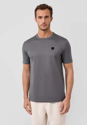 Man draagt een effen grijs T-shirt met korte mouwen en een klein zwart vierkant logo op de borst, gecombineerd met licht beige broeken tegen een grijze achtergrond.