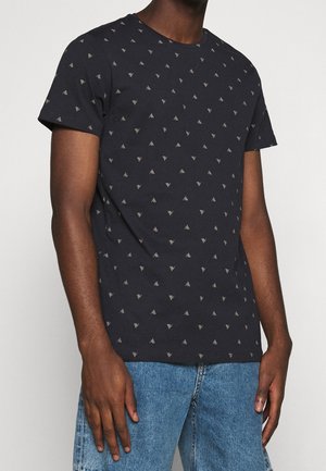 Man draagt een zwart T-shirt met korte mouwen met kleine grijze driehoekpatronen en een blauwe spijkerbroek, staand tegen een effen achtergrond.