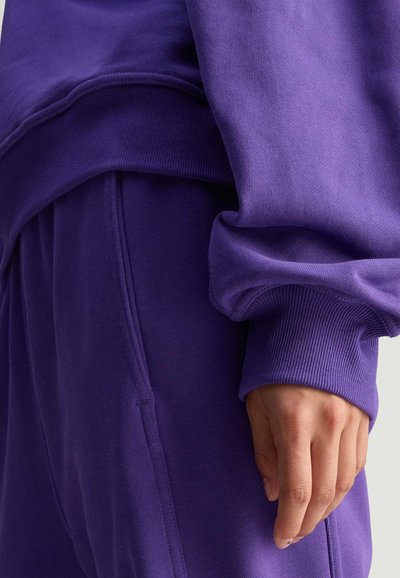 Persoon in een paarse sweatshirt en bijpassende paarse broek met geribde boorden, met een ontspannen hand naast zich tegen een lichte achtergrond.