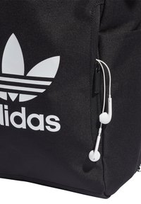 Svart ryggsäck med en framträdande vit Adidas-logotyp, texturerat tyg och vita hörlurar hängande från en liten öppning.