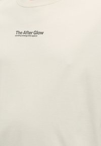 Πουκάμισο χρώματος κρέμα με μαύρο κείμενο στο στήθος που λέει «The After Glow στέλνει ενέργεια στο διάστημα.»