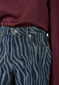 Jeans de mezclilla azul a rayas de cebra con cintura alta, que presentan un cierre de botón de metal y acentos dorados, combinados con una blusa texturizada burdeos.
