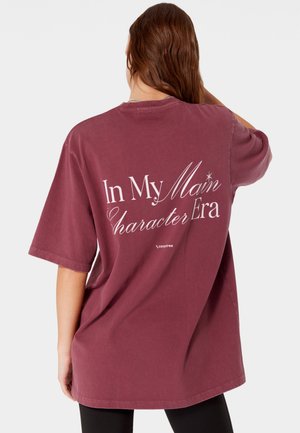 Osoba z długimi brązowymi włosami nosi oversize bordową koszulkę z białym napisem "In My Main Character Era" na plecach.