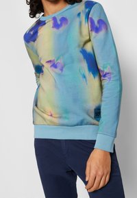 Felpa azzurro chiaro con motivi astratti multicolori, colletto e polsini a coste, con un piccolo patch logo sul lato inferiore.