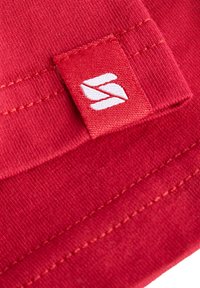 Roter Stoffpatch mit einem gestickten weißen Logo am Rand, feiner Textur und kontrastierenden Nähdetails.