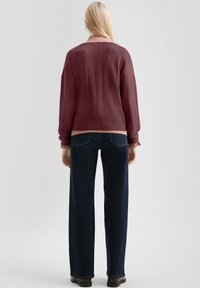 Maroonowy sweter o prążkowanej fakturze i okrągłym dekolcie, zestawiony z ciemnoniebieskimi prostymi spodniami i czarnymi butami. Wyświetlony widok z tyłu.