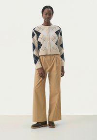 Sweter robiony na drutach w kolorze kremowym z granatowym wzorem argyle, okrągłym dekoltem i zapięciem na guziki. Połączony z beżowymi spodniami o szerokich nogawkach i ciemnymi butami.