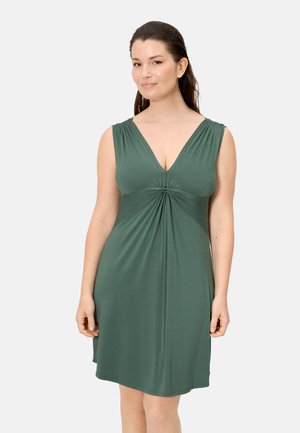 Mujer con cabello largo y oscuro, vestida con un vestido verde oscuro sin mangas hasta la rodilla, con detalle de tela reunida en el pecho, posando frente a un fondo liso.