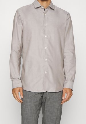 Mann trägt ein hellgraues, langärmliges Hemd mit Knöpfen und graue karierte Hose, steht vor einem weißen Hintergrund.