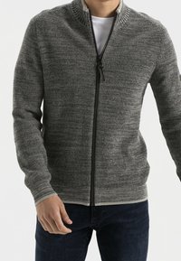 Harmaa vetoketjullinen neulepusero, jossa on ribbinen pinta, korkea kaulus ja kontrastivärinen vetoketju. Yhdistetty valkoiseen paitaan ja tummaan sinisiin farkkuihin.