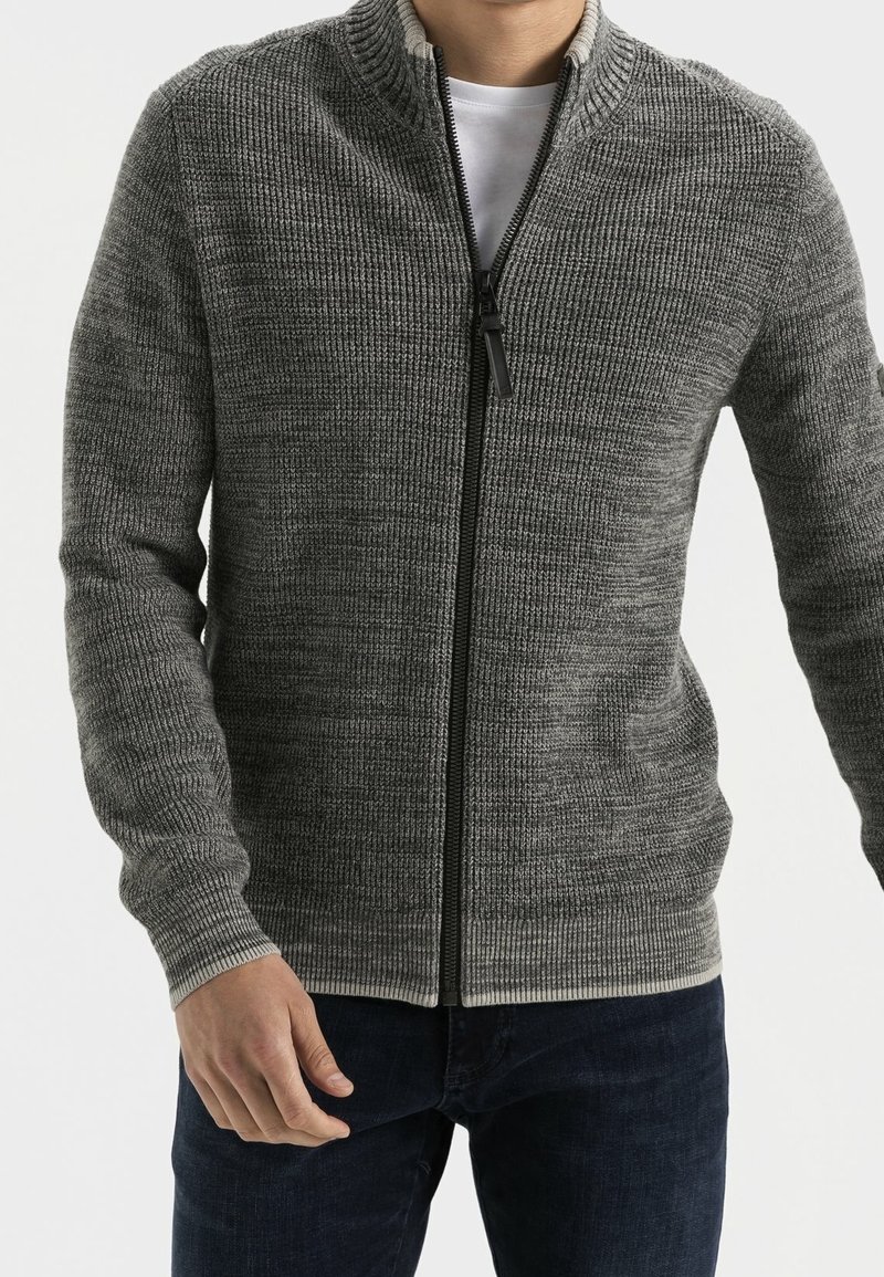 Harmaa vetoketjullinen neulepusero, jossa on ribbinen pinta, korkea kaulus ja kontrastivärinen vetoketju. Yhdistetty valkoiseen paitaan ja tummaan sinisiin farkkuihin.