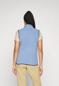 Person med langt flettet hår iført en lys blå fleece vest over en beige kortermet skjorte og beige bukser, vendt bort.