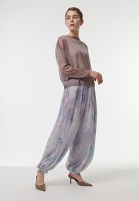 Lichtroze metallic trui met lange mouwen, gecombineerd met losse, gemusterde pastel palazzo-broeken. Het model draagt nude hoge hakken.