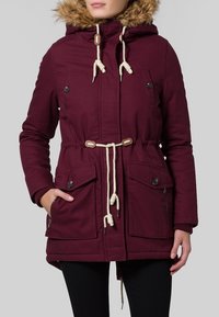 Mörkröd parka med en fuskpäls huva, dragsko i midjan, flera fickor och kontrasterande krämfärgade dragsnören. Slät textur.