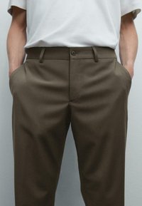 Man som bär olivgröna skräddarsydda byxor med händerna i fickorna och en instoppad vit t-shirt mot en enfärgad grå bakgrund.