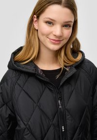 Junge Frau mit glattem blonden Haar, die eine schwarze, gesteppte Jacke mit Kapuze trägt und sanft vor einem schlichten Hintergrund lächelt.