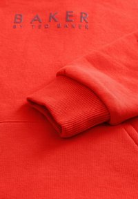 Sarkana džemperis ar ribotām aprocēm un teksturētu audumu. Zīmola logo "BAKER BY TED BAKER" ir izdrukāts tumšāk sarkanā krāsā.