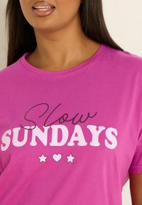 Vaaleanpunainen puuvillapaita, jossa on pyöreä kaula-aukko ja lyhyet hihat. Paidassa on teksti "Hitaat sunnuntait" valkoisena ja mustana, jota korostavat tähdet ja sydän.