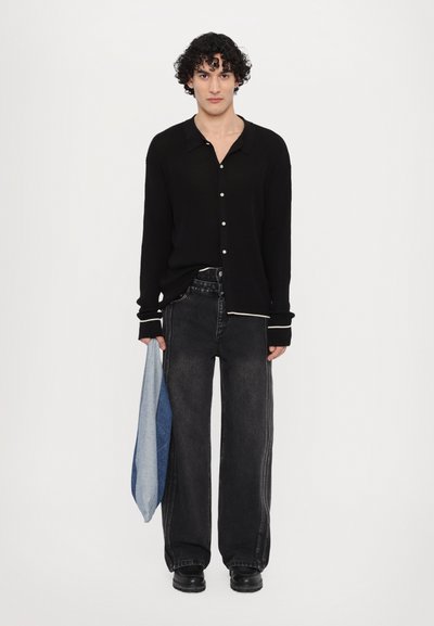 Camicia nera a coste con bottoni e colletto, abbinata a jeans in denim nero larghi con cuciture a contrasto. Tieni in mano un tessuto di denim blu piegato.