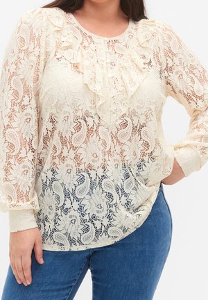 Femme portant un chemisier en dentelle crème à manches longues avec des motifs floraux et des volants, associé à un jean en denim bleu, posant avec une main sur la hanche.