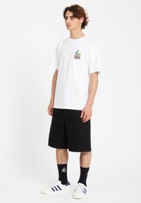 T-shirt en coton blanc avec un petit graphisme coloré sur la poitrine, associé à un short en denim noir et des baskets blanches avec des accents bleus.