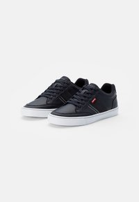 Sapatilhas de couro preto com solas de borracha branca, apresentando um design de atacador, riscas laterais contrastantes e detalhes de marca na língua.