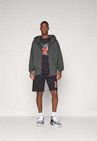 Felpa con zip grigia scura con logo, t-shirt nera con grafica, pantaloni corti neri con la lettera rossa B e sneaker grigie con dettagli neri.