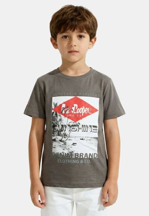 Garçon aux cheveux bruns portant un t-shirt gris avec un motif de plage et un pantalon blanc, debout devant un fond clair uni.