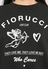 Camisa preta com texto branco "FIORUCCI LOVE CLUB," um cupido apontando uma flecha, um coração, e as frases "Eles gostam de mim, não gostam" e "Quem se importa."
