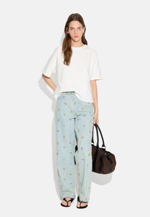 Kobieta ubrana w luźną białą bluzkę z krótkim rękawem, jasnoniebieskie jeansy z szerokimi nogawkami i drobnym kwiatowym wzorem, czarne sandały, trzyma brązową i czarną torebkę.