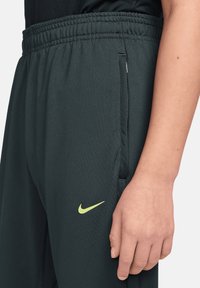 Dunkelgrüne Sporthose aus leichtem Stoff mit elastischem Bund, Seitentaschen und kleinem gelbem Nike-Logo.
