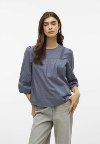 Modrá bavlněná blůza s nabíranými rukávy, jemnou tečkovanou texturou a krajkovými akcenty na ramenou. V kombinaci se světlými šedými kalhotami s vysokým pasem.