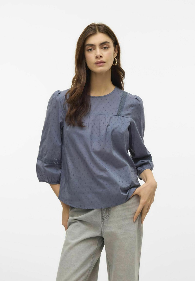 Modrá bavlněná blůza s nabíranými rukávy, jemnou tečkovanou texturou a krajkovými akcenty na ramenou. V kombinaci se světlými šedými kalhotami s vysokým pasem.