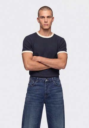 Ung mand med kort hår står med armene over kors iført en sort t-shirt med hvid kant og blå jeans mod en hvid baggrund.